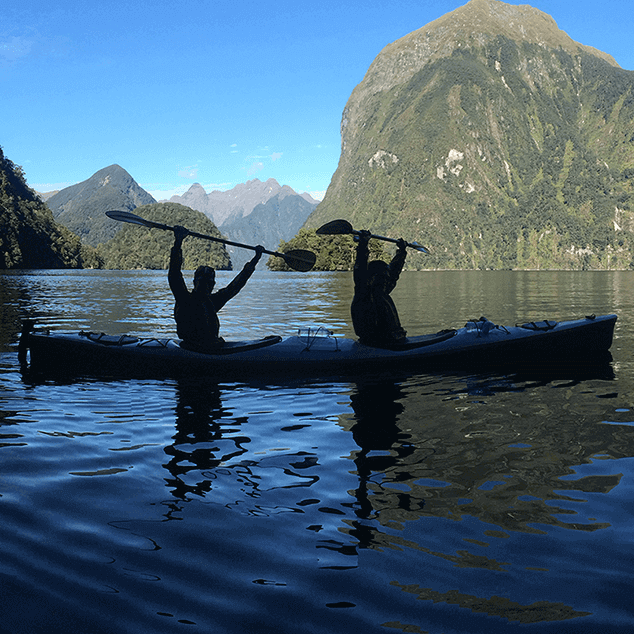 Kayaker lifting paddle above his head with mountains in the background
