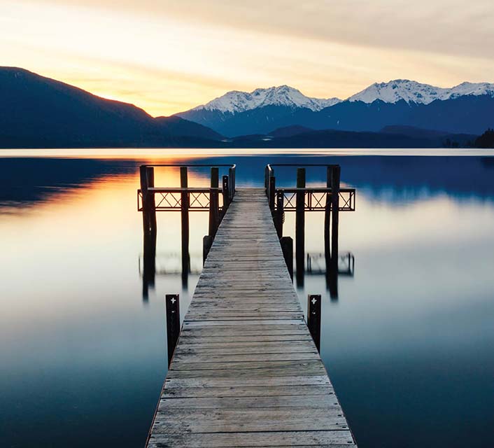 Marakura Yacht Club wharf reaching out into Lake Te Anau on calm waters with snow capped mountains and the orange colours of sunset reflecting upon the water 