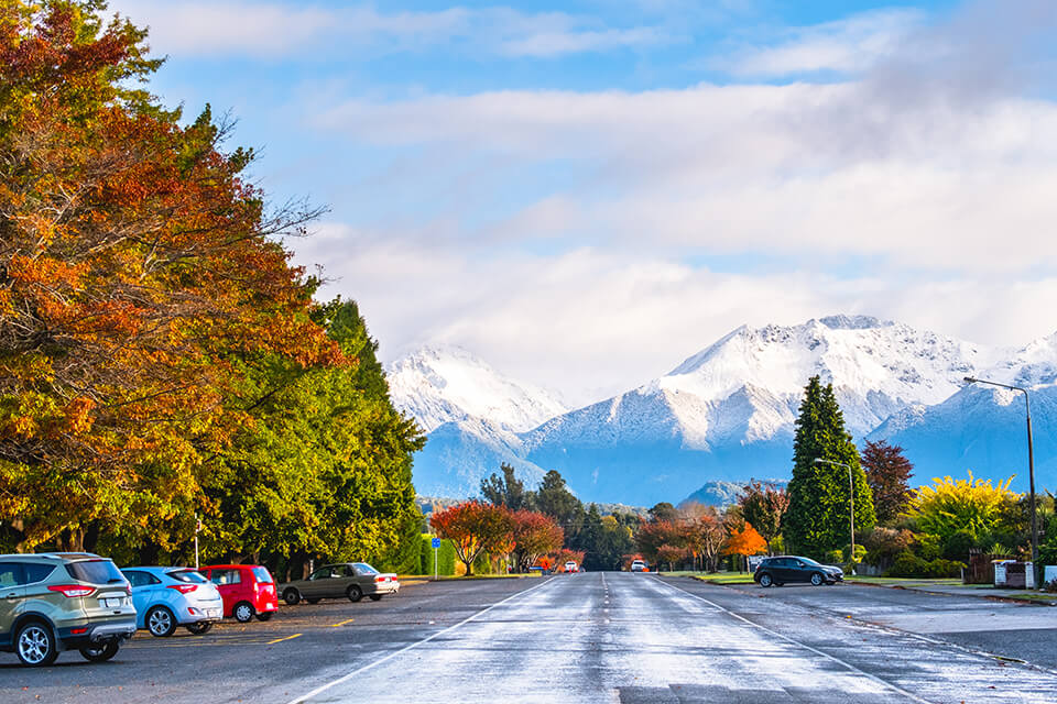 Looking down a long straight road bordered with Autumnal coloured trees, towards mountains with snowy tops